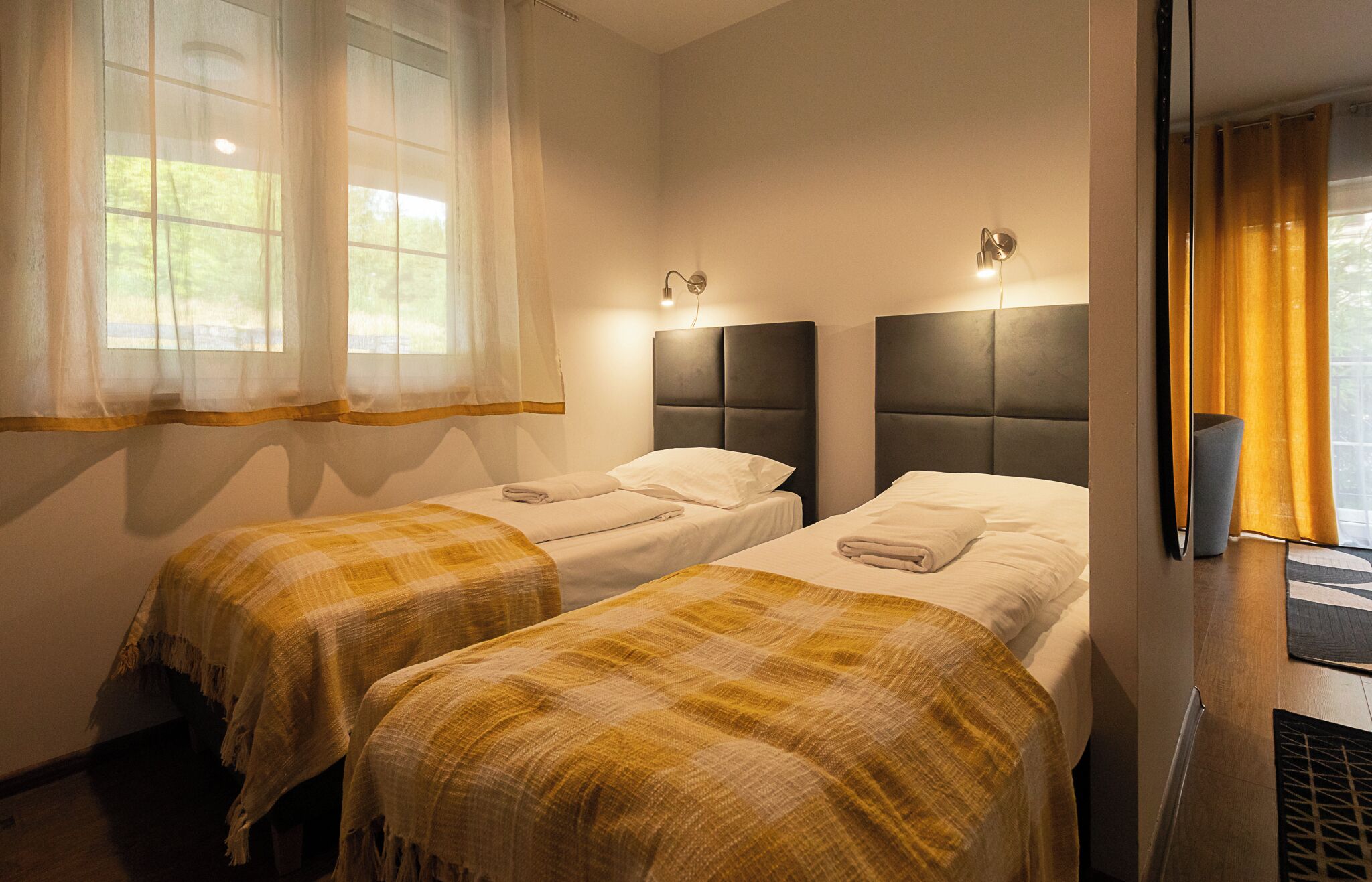 Quarto de hotel com duas camas, cada uma com cabeceira, uma janela com cortinas translúcidas e luminária fixada na parede.