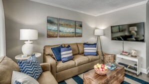 Ein Wohnzimmer mit einem beigen Sofa, blau-weißen Streifen-Kissen, einem Couchtisch mit Obst, einem weißen Beistelltisch mit einer Lampe und einem an der Wand montierten Flachbildfernseher.