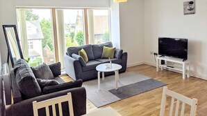 A living room with a sofa, a coffee table, a TV, and a large window. - Nidus Flat (Cambridge)