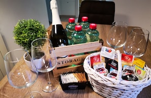 A wooden table with a bottle of champagne, several green bottles, empty wine glasses, a small potted plant, and a basket containing various snacks and a box of chocolates. - Casa Da Ponte (Koper)