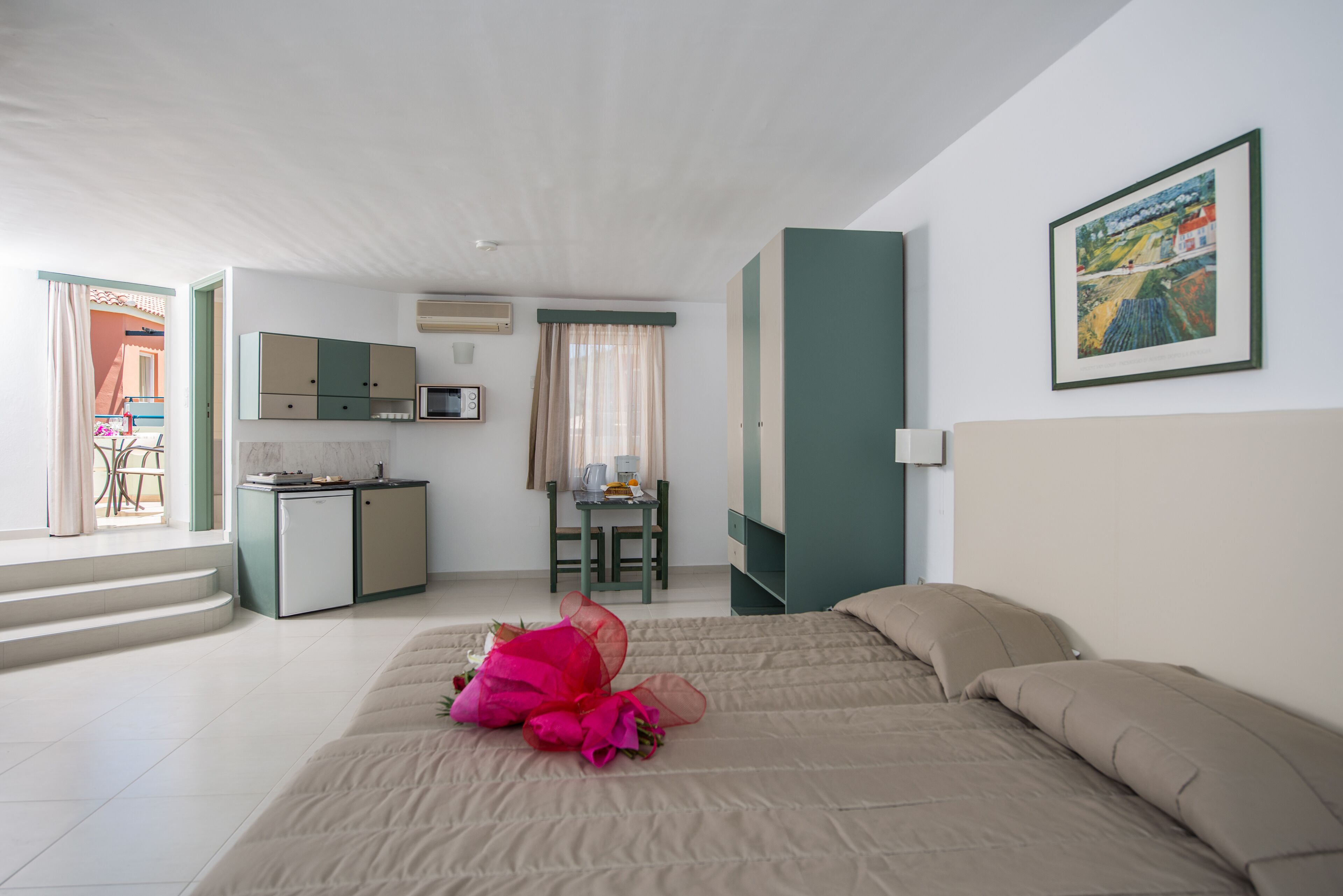 A hotel room with a bed, a desk, a kitchenette, and a view of the outside.