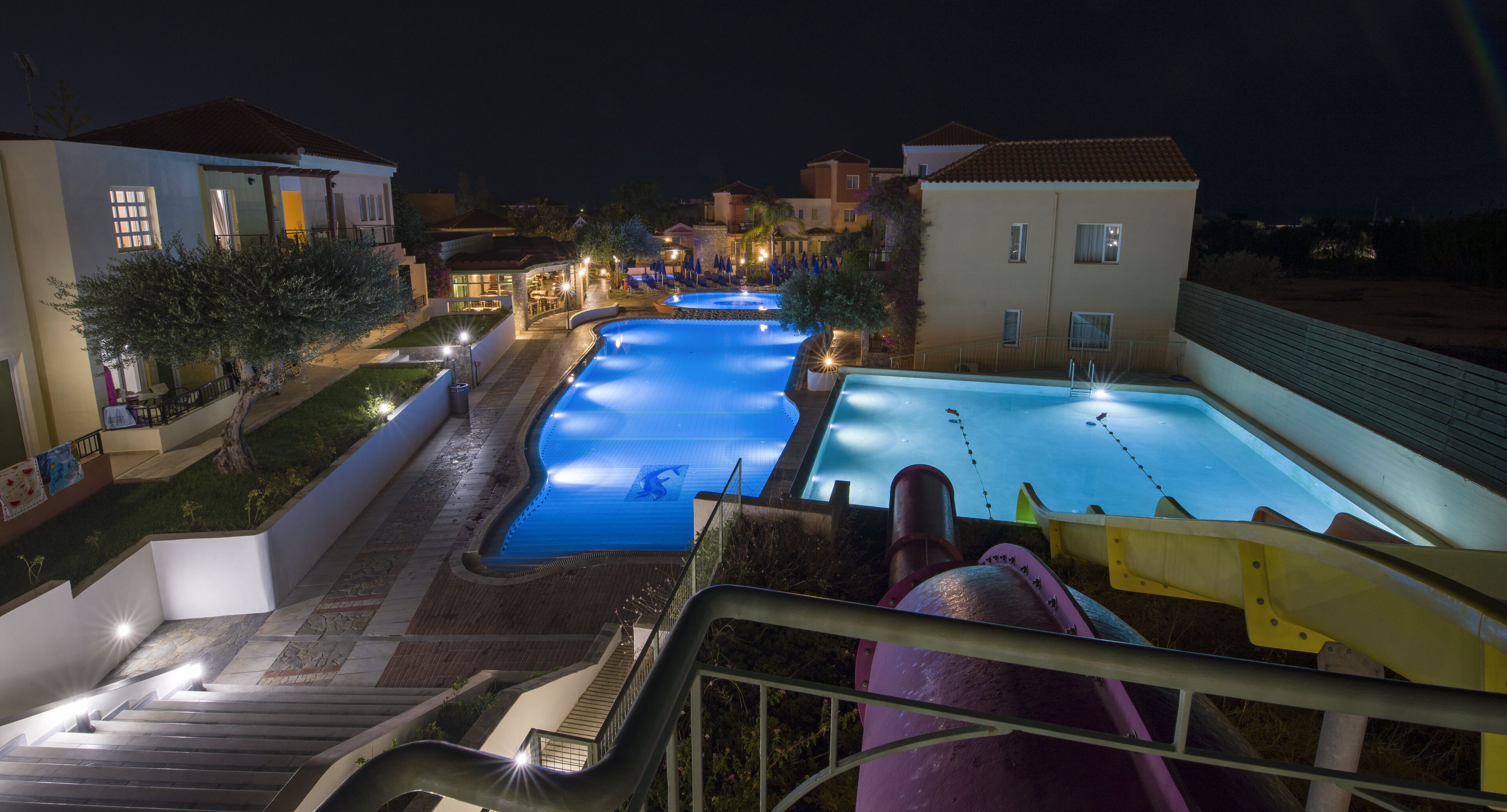 A nighttime view of a resort with multiple swimming pools, a slide, and a staircase leading to the pools.