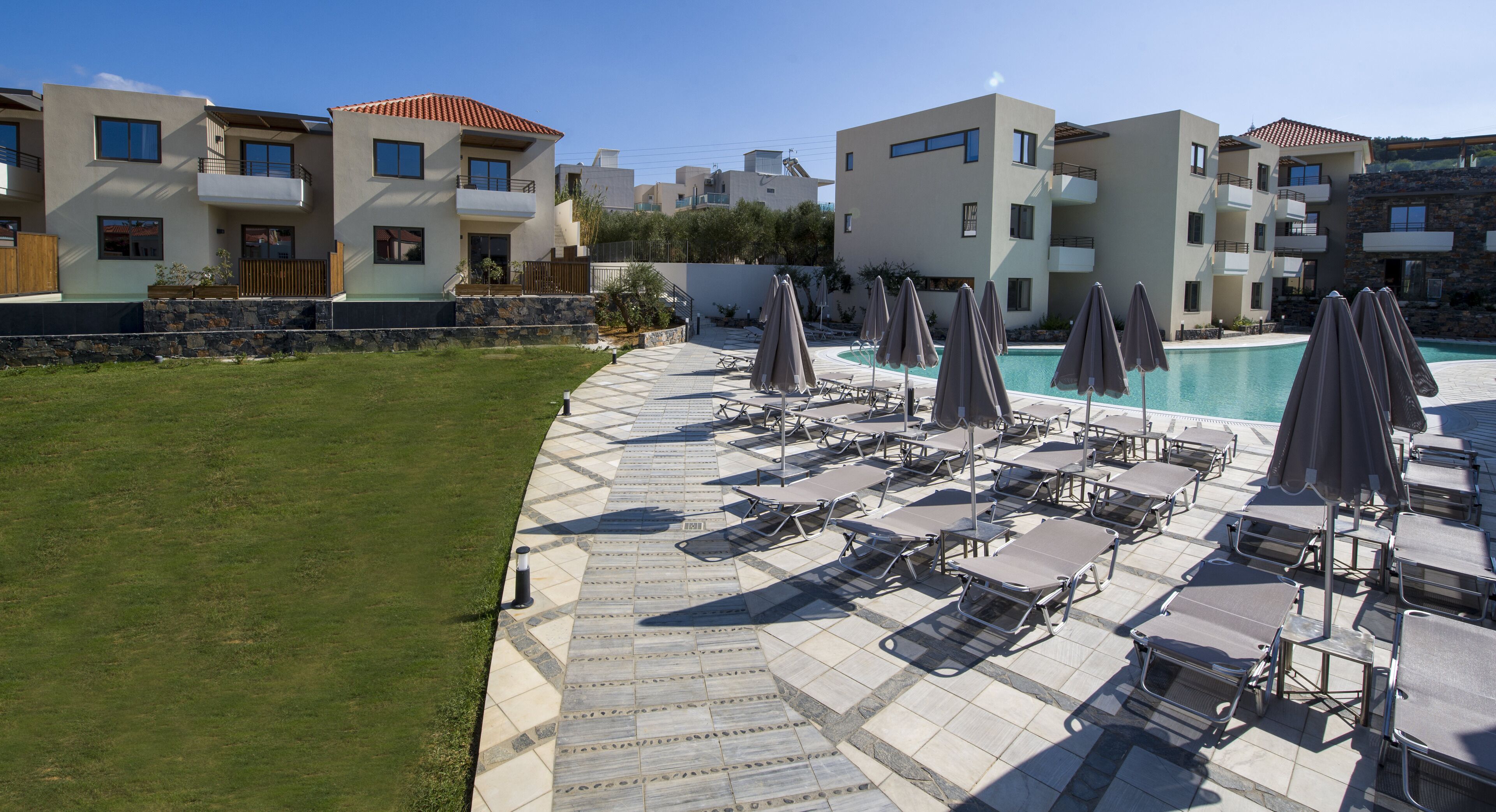 A poolside area with lounge chairs and umbrellas, a stone wall, and modern buildings in the background.