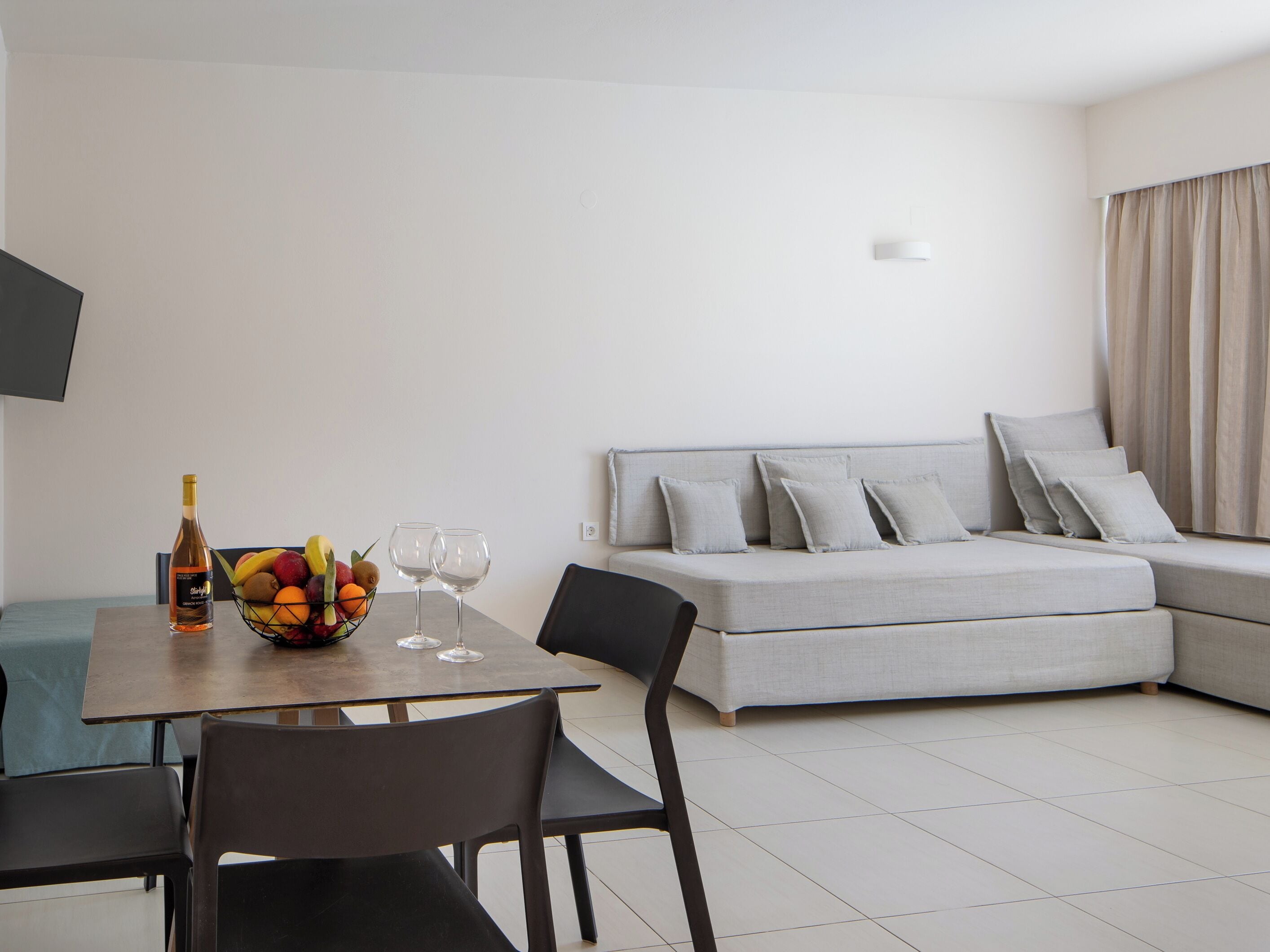 A modern living room with a sofa, a dining table with chairs, a fruit bowl, and a bottle of wine.