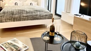 A modern hotel room with a large bed, a bedside table with a bottle and glasses, a small round table, a magazine, and a TV on a stand. - Grace-Design-Apartment Deluxe in the center of Munich, top location (München)