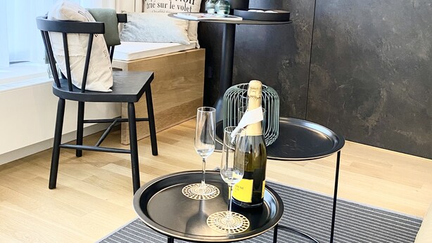 A modern room with a dining table, a chair, a small side table with a bottle of champagne, and a wall-mounted light fixture. - Grace-Design-Apartment Deluxe in the center of Munich, top location (München)