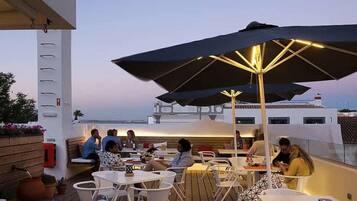 An outdoor dining area with tables and chairs, umbrellas, and a view of a boat in the background.