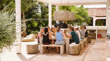 Un patio al aire libre con personas sentadas en mesas, rodeado de follaje y una estructura de pérgola.