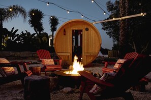 Une structure en bois dotée d’une porte circulaire, entourée d’un espace extérieur aménagé avec des chaises rouges et un foyer.