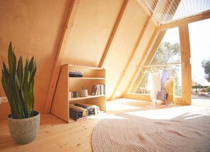 A cozy attic bedroom with wooden walls, a large window, a bookshelf, and a potted plant.