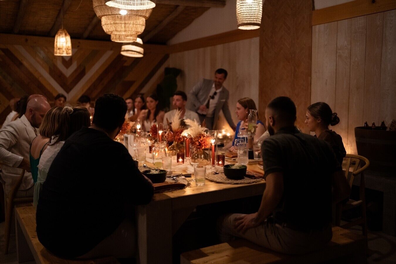 A group of people seated around a wooden table with candles and drinks, in a room with wooden walls and hanging lights.
