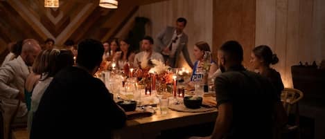 Um grupo de pessoas sentadas ao redor de uma mesa de madeira com velas e bebidas, em um ambiente com paredes de madeira e luminárias pendentes.