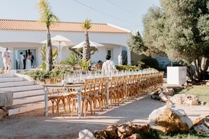 Un espace extérieur aménagé pour les repas, avec des tables et des chaises, des palmiers et un bâtiment en arrière-plan.