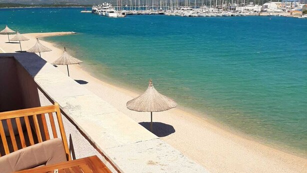 A wooden deck with a lounge chair and a small table, overlooking a beach with clear turquoise water and a marina in the background. - Apartment with sea view and next to the beach (20m) (Betina)