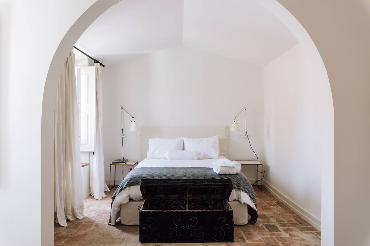 A bedroom with a large bed, a bench, and a window with curtains.