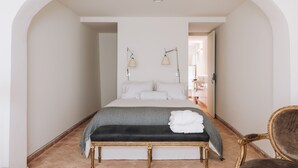 A bedroom with a large bed, a bench, and a chair. - Travassos 11 (Elvas)
