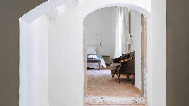 An arched doorway leading to a bedroom with a bed, a chair, and a lamp. - Travassos 11 (Elvas)