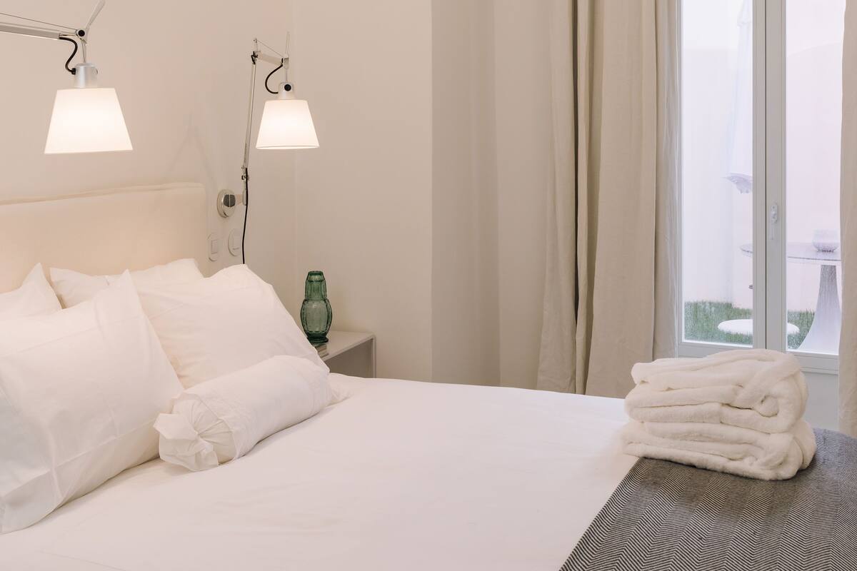 A hotel room with a bed, two bedside lamps, a nightstand with a green glass, and a window with curtains.