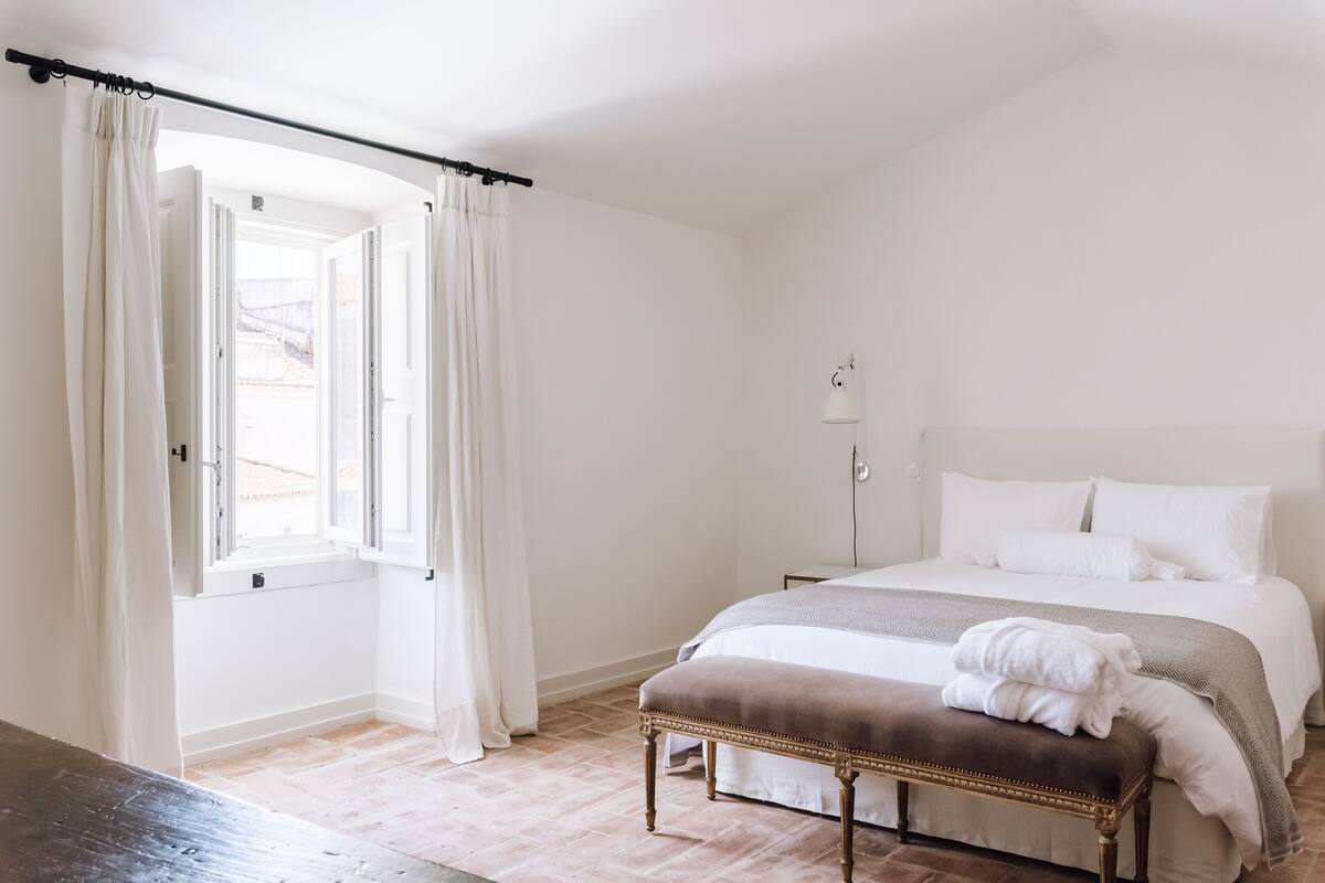 A bedroom with a bed, a bench, a lamp, and a window with curtains.