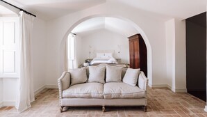 A room with a large arched doorway leading to a bedroom, a beige sofa in the foreground, and a wooden cabinet in the background. - Travassos 11 (Elvas)