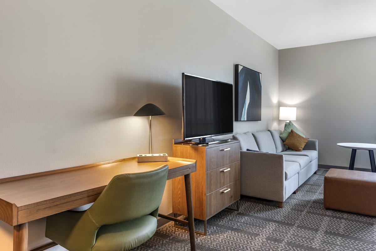 A modern hotel room with a wooden desk, a green chair, a sofa, a TV, and a lamp.
