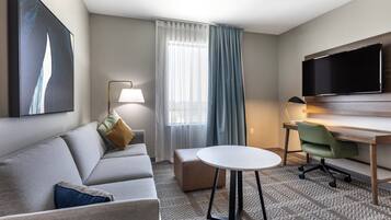 A modern hotel room with a grey sofa, a small round table, a green office chair, a flat-screen TV mounted on the wall, and a large window with sheer curtains.