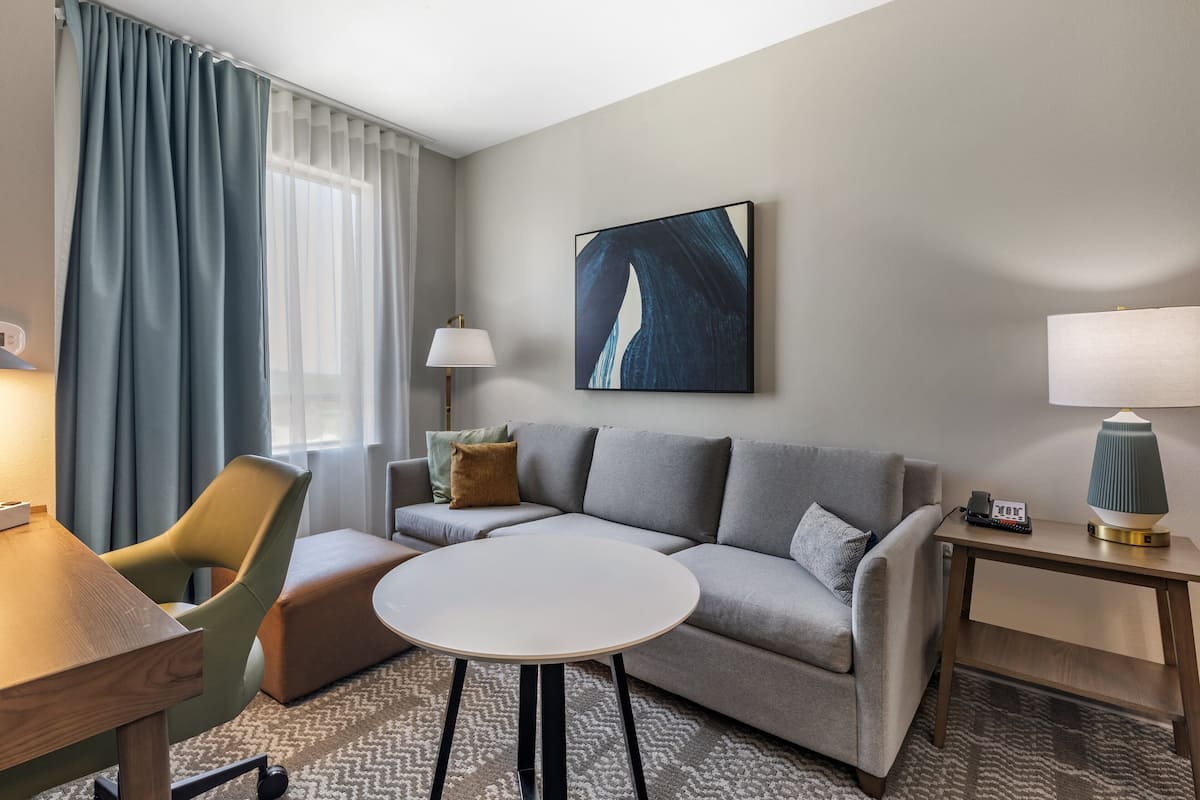 A modern hotel room with a grey sofa, a round table, a wooden side table, a lamp, and a painting on the wall.