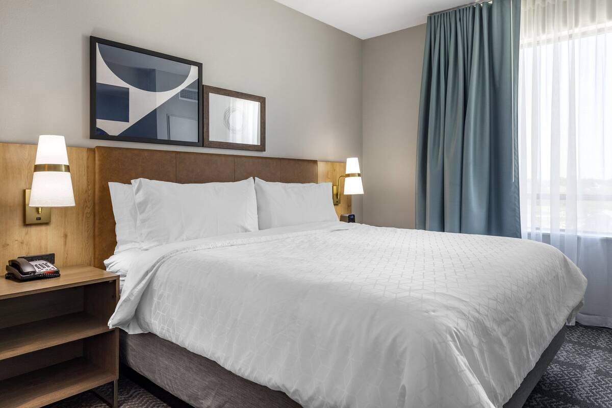 A hotel room with a large bed, two bedside lamps, a telephone on the nightstand, and a window with curtains.