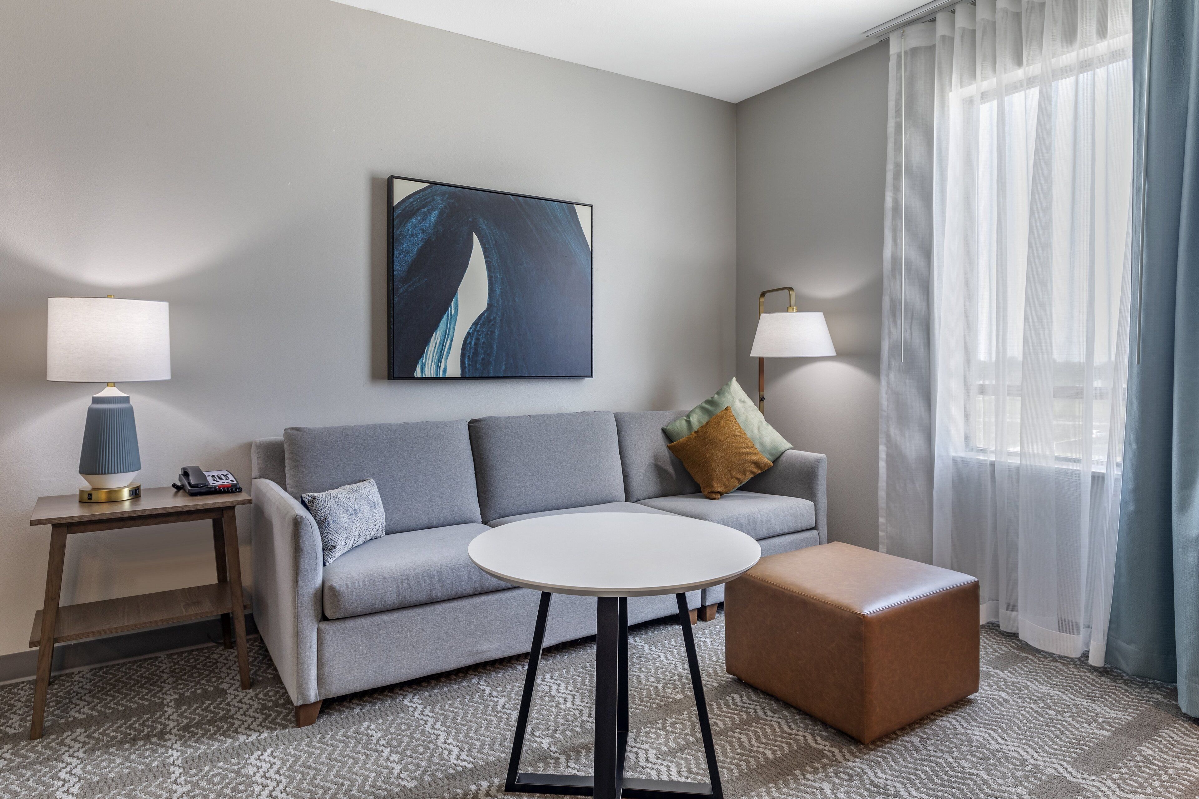 A modern hotel room with a grey sofa, a round white table, a brown ottoman, and a large abstract painting on the wall.