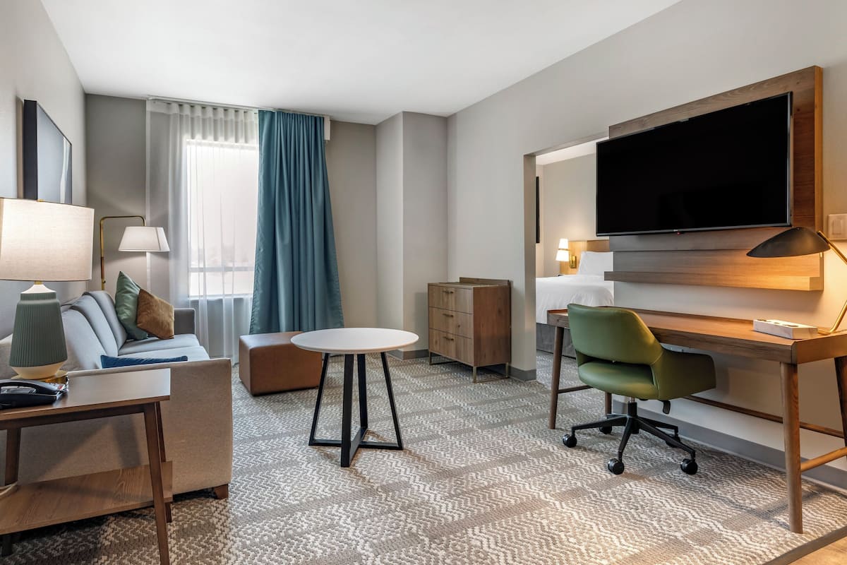 A hotel room with a sofa, a small round table, a desk with a chair, a television, and a wooden cabinet.