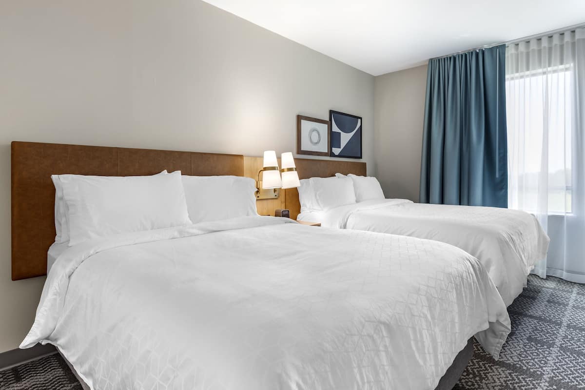 A hotel room with two beds, a headboard, bedside lamps, a framed picture, and a window with curtains.