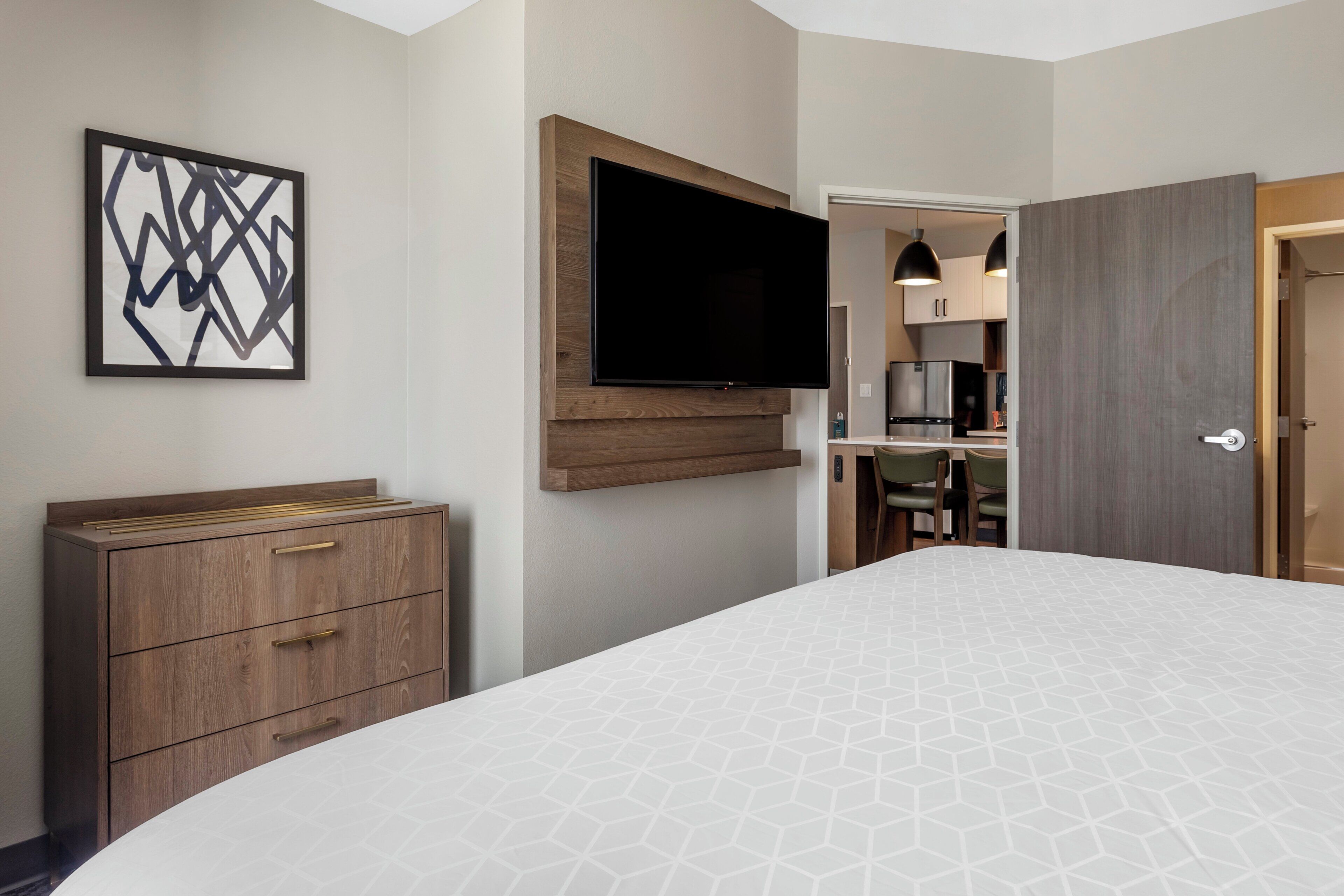 Une chambre d’hôtel comprenant un lit, une commode, un téléviseur à écran plat fixé au mur et un coin repas visible à travers une porte ouverte.