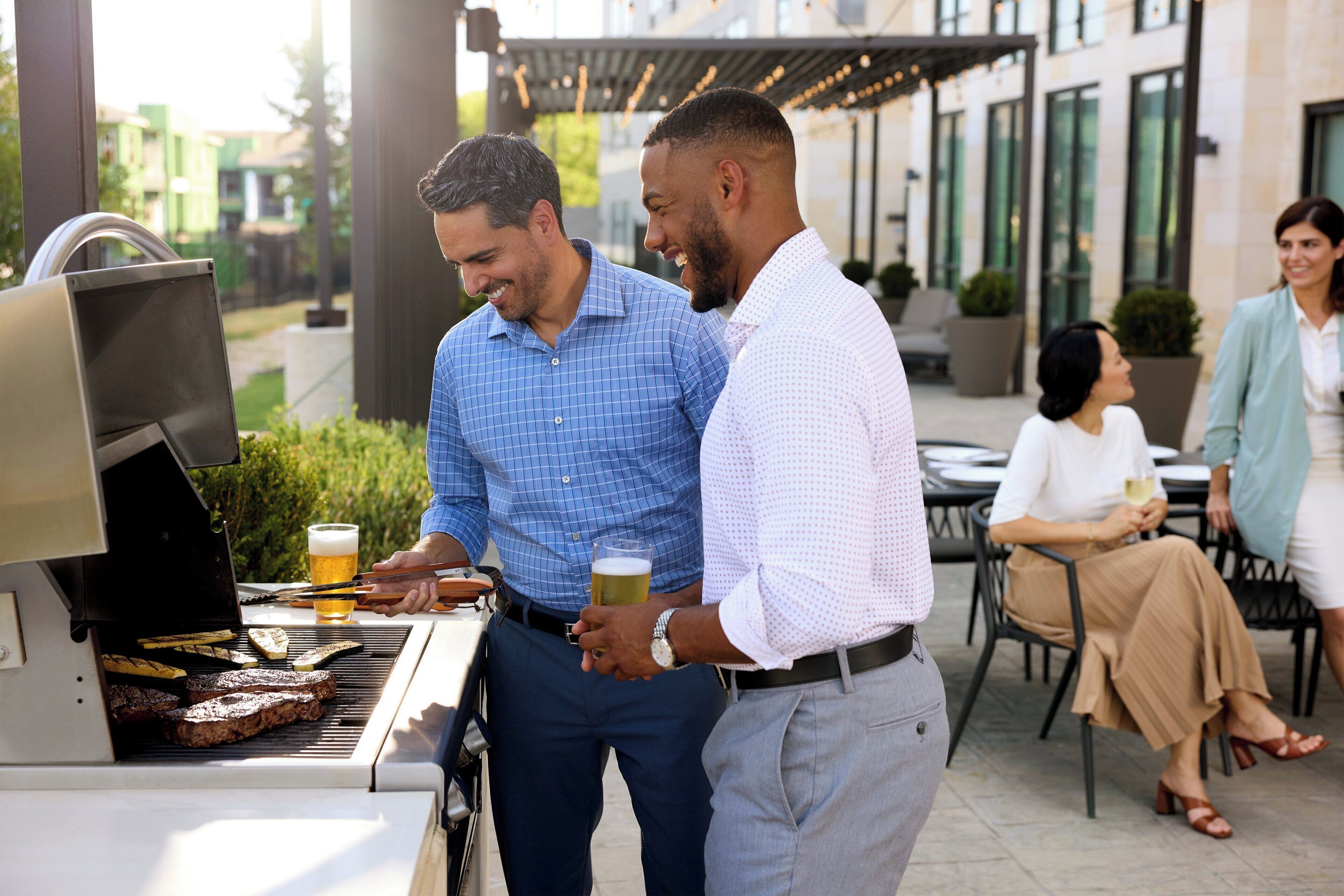 A man grilling steaks outdoors with a beer in hand, another man observing, and a woman seated nearby.