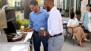 Un homme est en train de faire griller des steaks à l’extérieur, une bière à la main, un autre homme l’observe, et une femme est assise à proximité.
