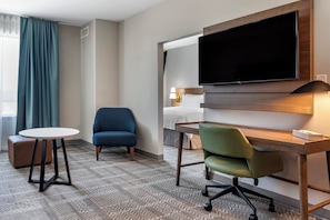 Quarto de hotel com uma cama, televisão na parede, escrivaninha com cadeira, mesa redonda pequena e um sofá.