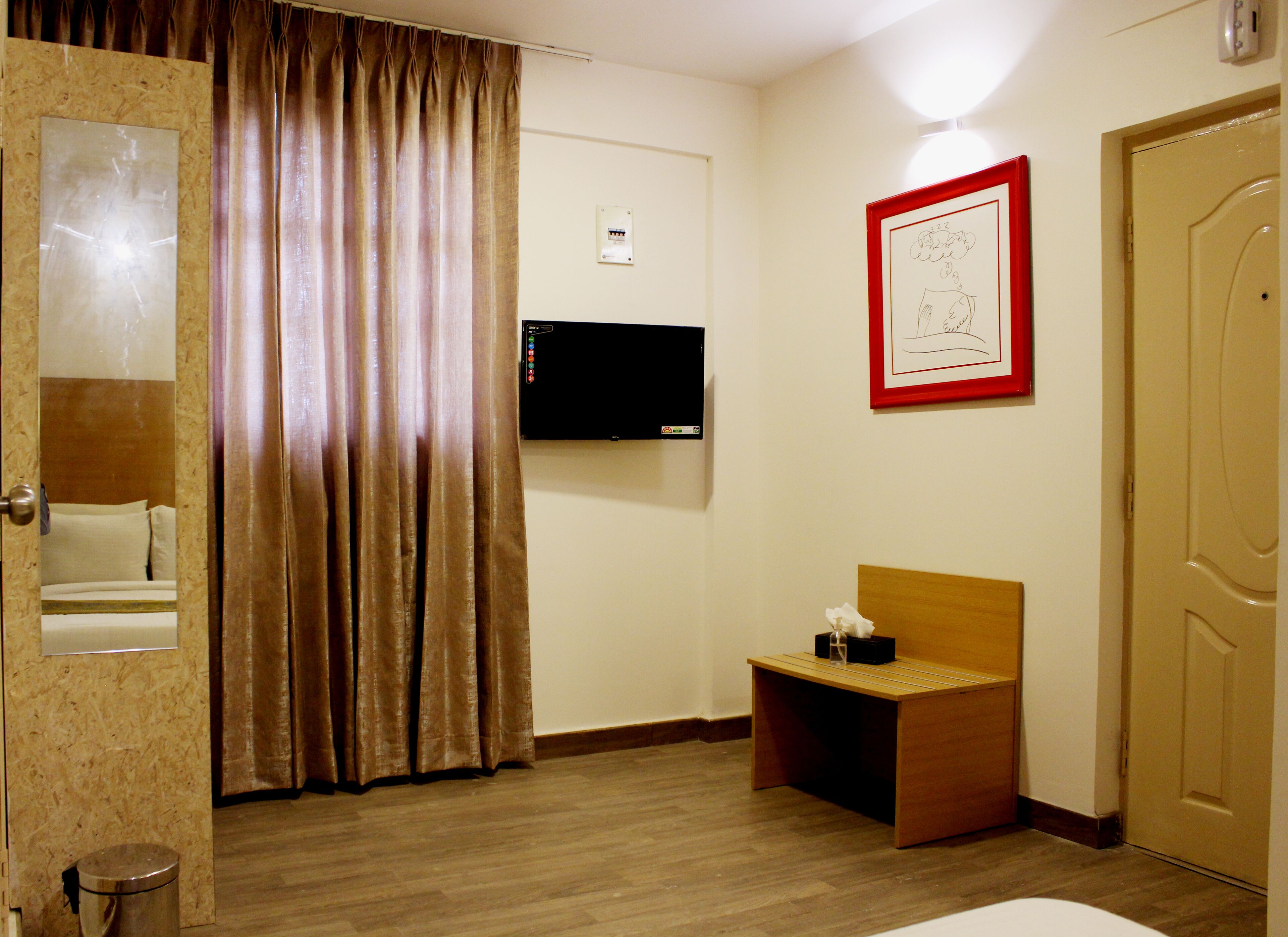 A hotel room with a wooden floor, a bed with a headboard, a bedside table with a tissue box, a wall-mounted TV, a framed picture, and a door.