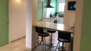A compact kitchen with a wooden table, two bar stools, a TV mounted on the wall, and a green door.