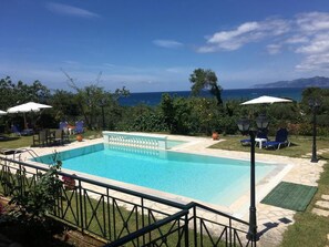 A rectangular swimming pool with a tiled deck, surrounded by lounge chairs and umbrellas, with a view of the sea and mountains in the background. - Pension Skala (Corfu)