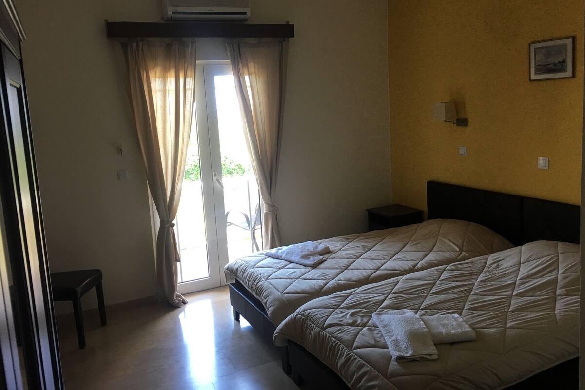 A bedroom with two beds, a window with curtains, an air conditioner above the window, and a small bedside table with a lamp.