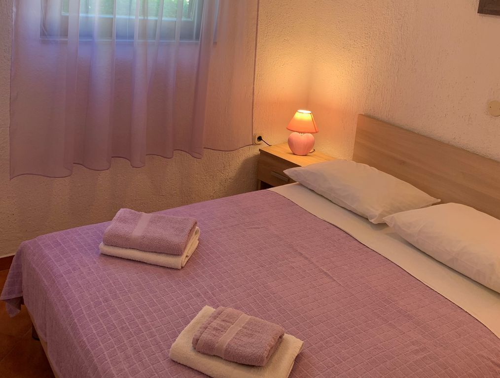 Une chambre à coucher avec un lit, deux oreillers, une table de chevet avec une lampe et une fenêtre avec des rideaux légers.