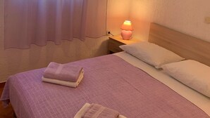 Un dormitorio con cama, dos almohadas, una mesita de noche con lámpara y ventana con cortinas traslúcidas.