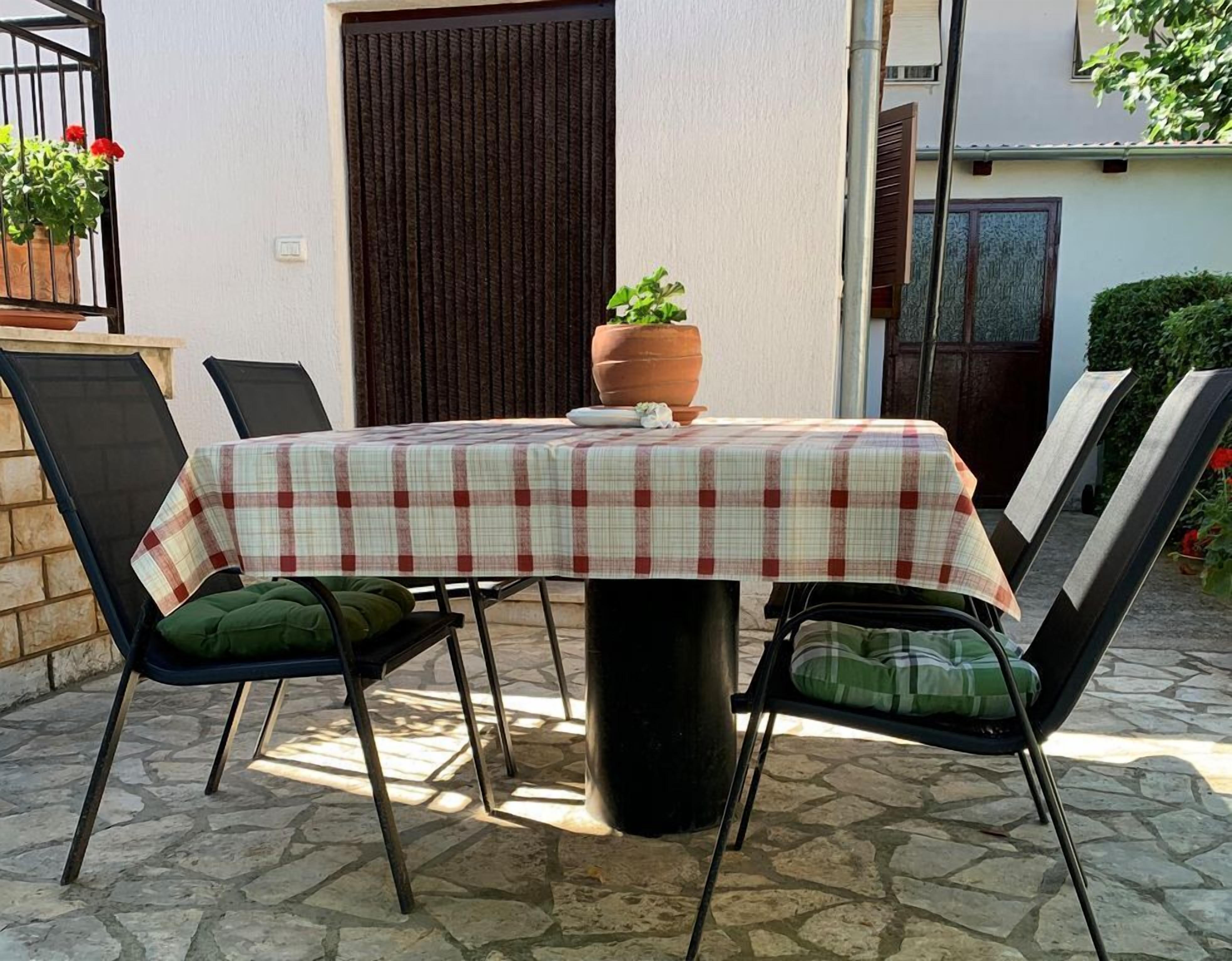 Une terrasse extérieure avec une table recouverte d’une nappe à carreaux, entourée de chaises noires dotées de coussins verts.
