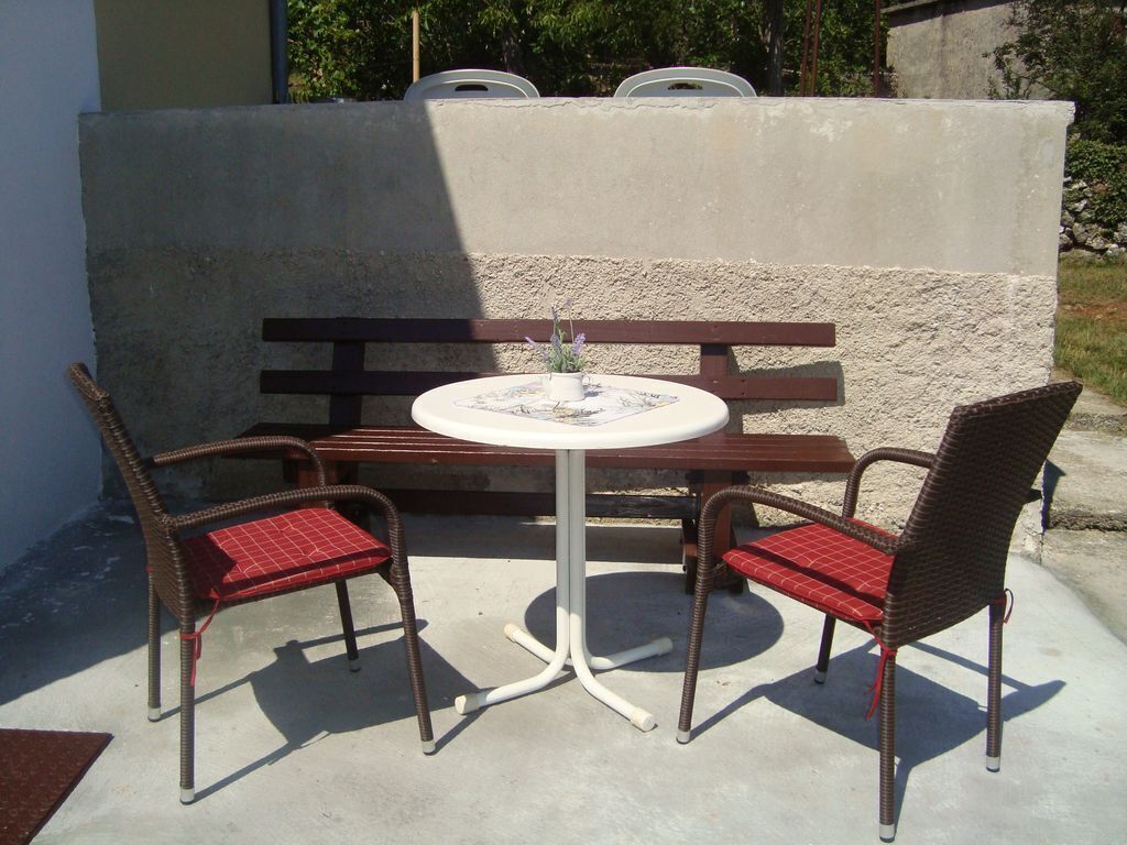 Een terras met een ronde tafel, twee rieten stoelen met rode kussens en een houten bank.