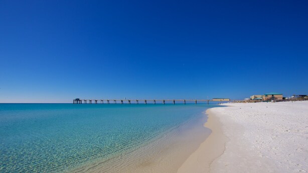 Una playa con un muelle que se adentra en el agua, cielos despejados y una orilla arenosa.