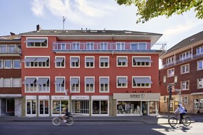 A multi-story building with a red facade, storefronts on the ground floor, and a balcony on the second floor. - Central Hotel Münster (Münster)