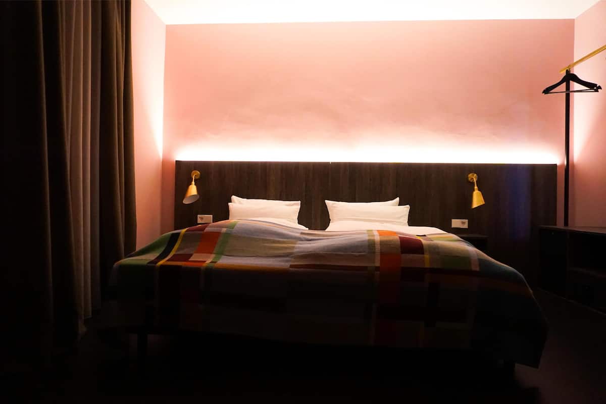 A hotel room with a bed, a nightstand, a lamp, and a wall-mounted light.