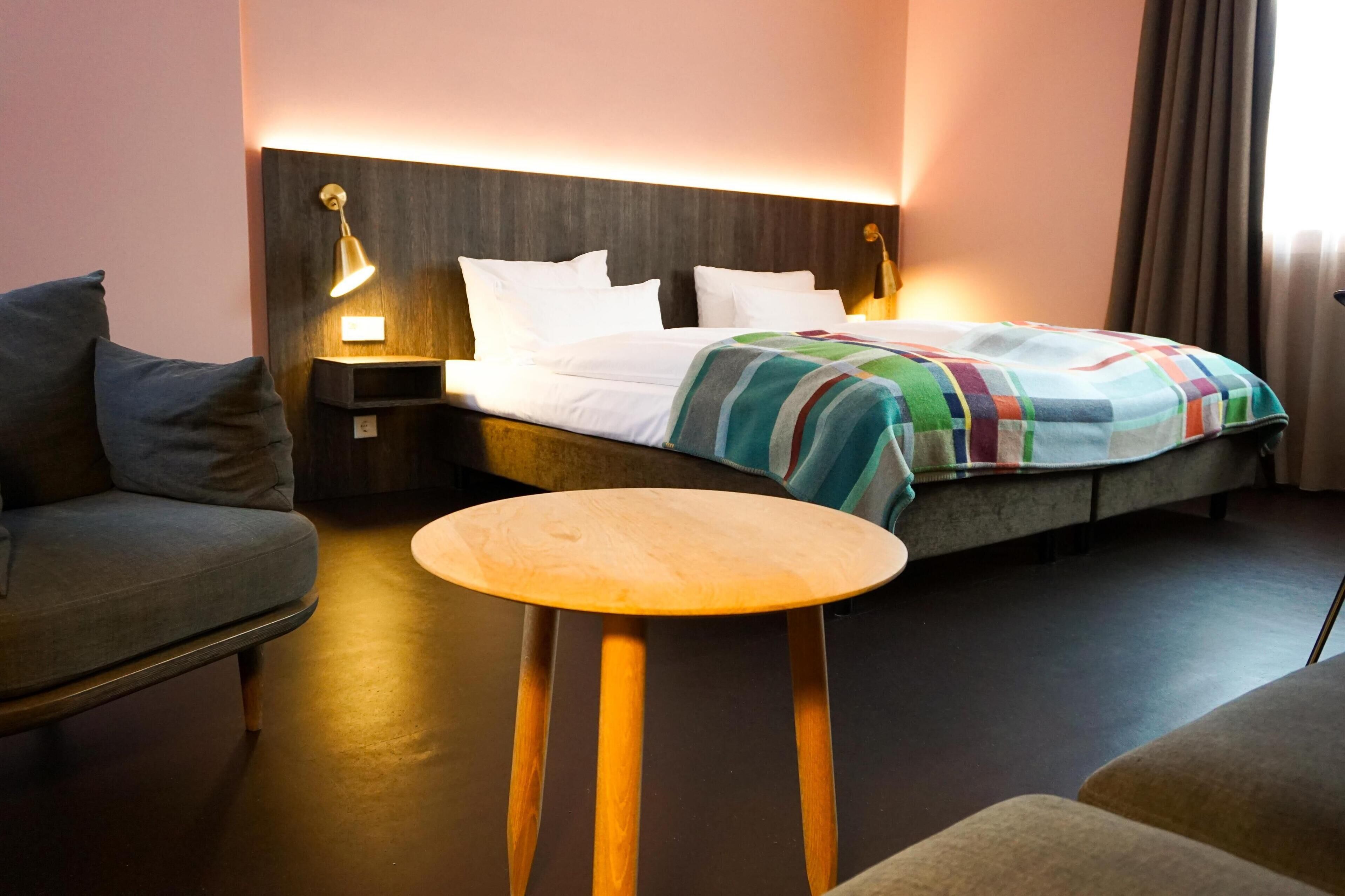 A hotel room with a bed, a bedside table, a lamp, and a sofa.
