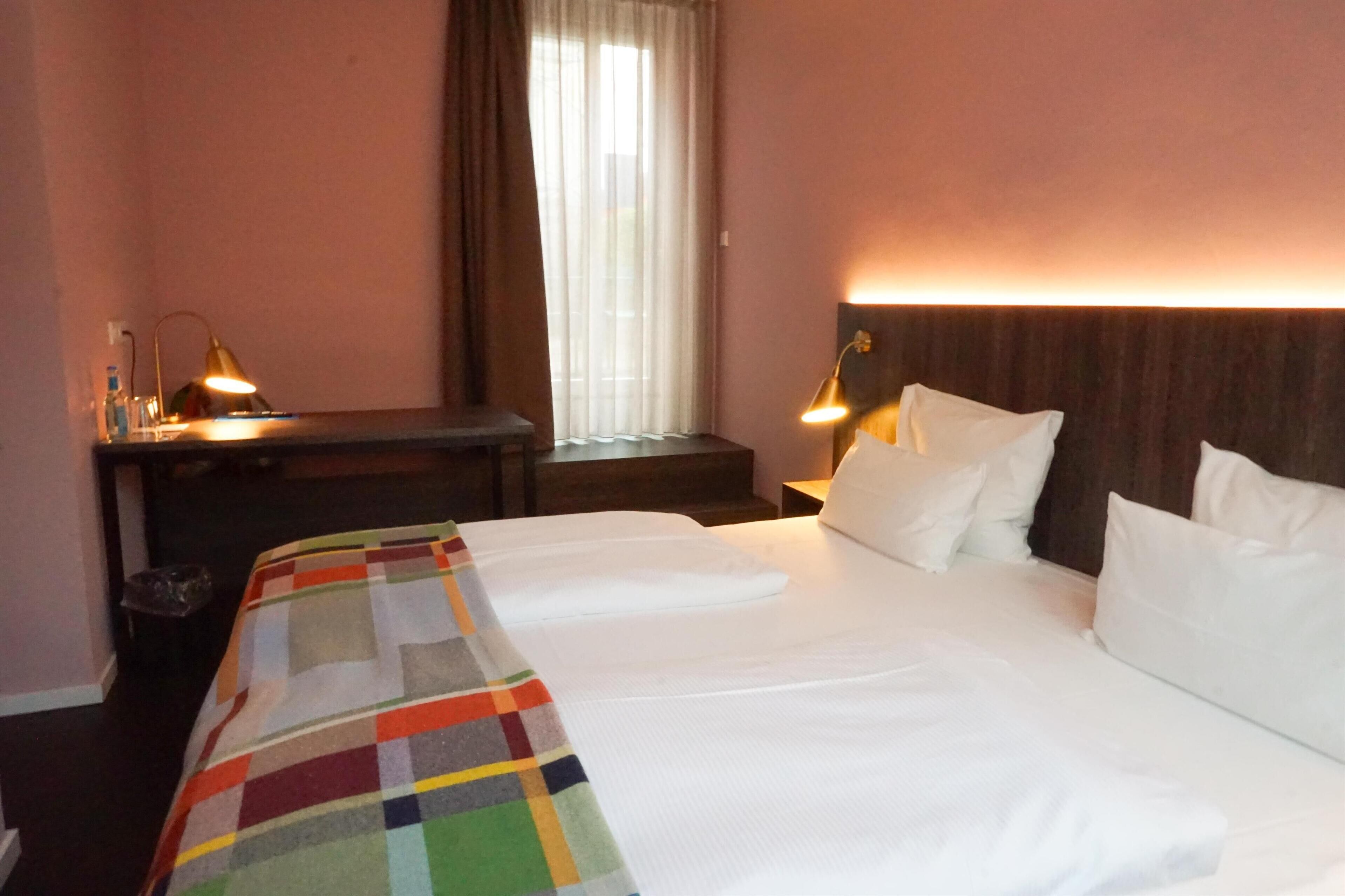 A hotel room with a large bed, two bedside tables with lamps, a window with curtains, and a colorful throw blanket.