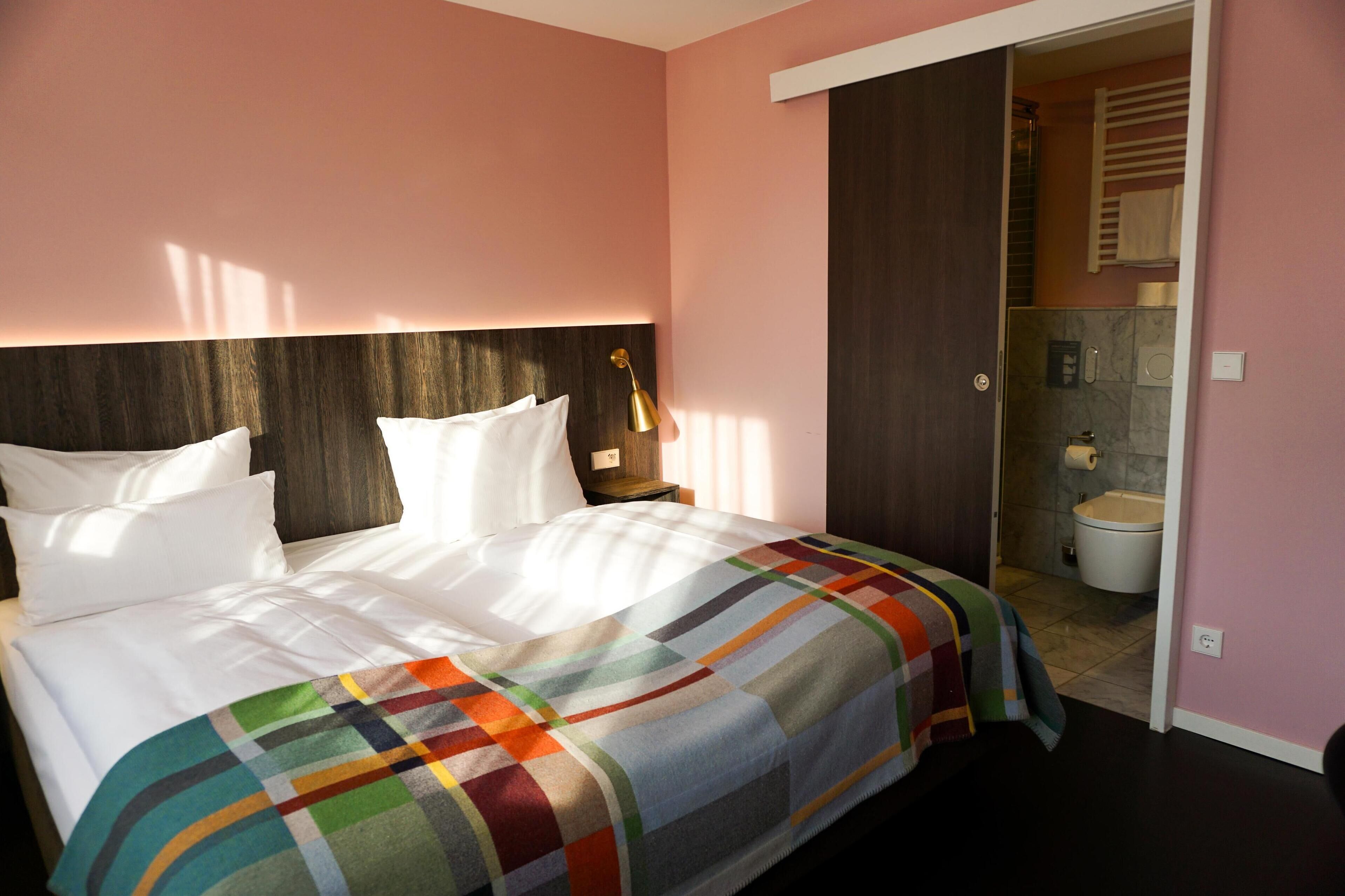 Habitación de hotel con cama, mesita de noche, lámpara y vista al baño.
