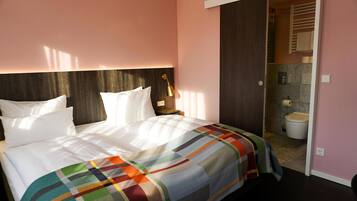 Habitación de hotel con cama, mesita de noche, lámpara y vista al baño.